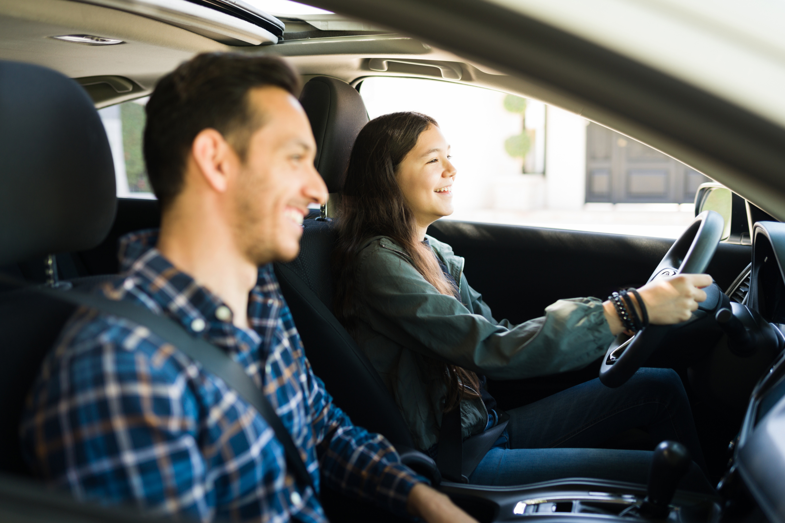 Happy handsome dad and teen daughter laughing while driving. Adolescent girl practicing her driving skills with her father